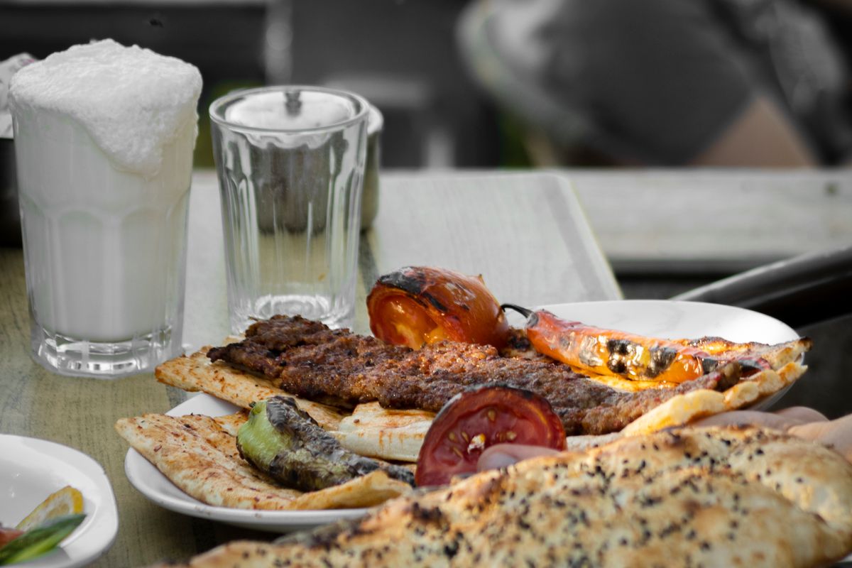Grilled Turkish kebab on flatbread with charred tomato and pepper, a glass of ayran and pide bread